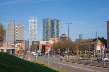 Manchester 'ın gündüz manzarasında Deansgate Meydanı kuleleri, tarihi tuğla binalar, trafik işaretleriyle dolu yoğun bir cadde ve çimenli bir set var..