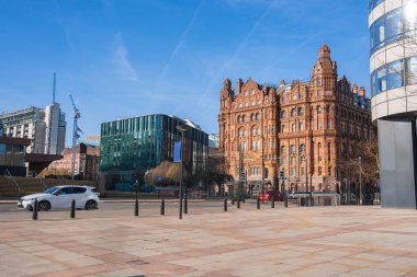 Manchester 'ın tarihi Midland Oteli manzarası, modern cam bir bina, arabaları ve yayaları olan temiz bir plaza, ve açık mavi bir gökyüzü.