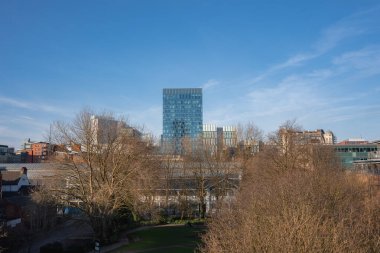 Manchester manzarasında modern cam gökdelen, tarihi ve çağdaş binalar ve açık mavi gökyüzünün altında çıplak ağaçlar olan bir park var..