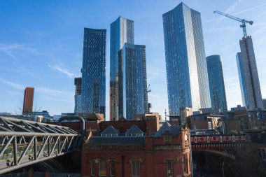 Manchester, İngiltere manzarası, Deansgate Meydanı gökdelenlerini gösteriyor, kırmızı tuğlalı Victoria binası, yaya köprüsü, ve bir vinç..