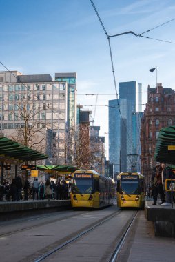 Manchester istasyonunda yolcu taşıyan iki sarı Metrolink tramvayı. Beetham Kulesi ve kırmızı tuğlalar açık mavi gökyüzünün altında duruyor..