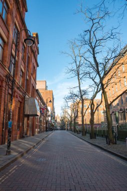 Manchester, İngiltere 'de kırmızı tuğlalı bir cadde, kırmızı tuğlalı binalar, çıplak ağaçlar ve temiz bir gökyüzü altında modern ve tarihi mimarinin bir karışımı..