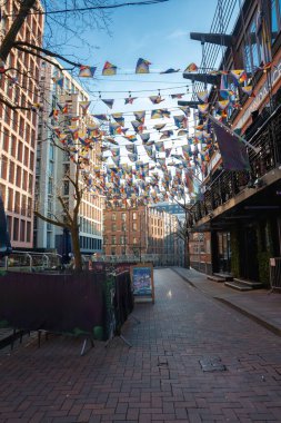Canlı bir Manchester caddesi, kırmızı tuğlalı kaldırım, modern ve tarihi binalar, üzerinde renkli bayraklar, ve açık mavi gökyüzünün altında açık hava koltukları..