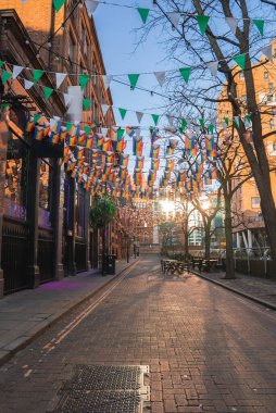 Manchester, İngiltere 'de renkli kiraz kuşu, tarihi tuğla binalar, çıplak ağaçlar, açık hava oturma yerleri ve arka planda yapıya benzer bir kilise içeren bir cadde..