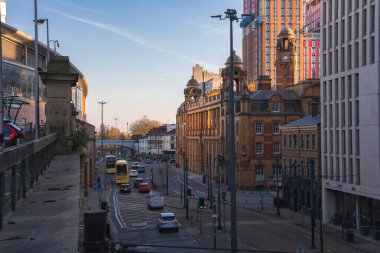 Manchester, İngiltere 'de canlı bir cadde, kırmızı tuğlalı bina, modern cam yapı, sarı çift katlı otobüs, ve inşaat halindeki bir gökdelen.