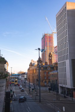 Manchester sokak sahnesinde saat kulesi olan kırmızı tuğlalı bir bina, inşa halinde olan modern bir gökdelen ve sarı çift katlı bir otobüs var..