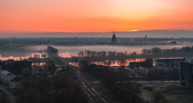 Riga, Letonya üzerinde sakin bir gün doğumu, Aziz Peter Kilisesi kulesi, Daugava Nehri 'ni kaplayan sis, bir demiryolu köprüsü ve yumuşak ışık altında kentsel yapılar.