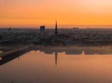 Riga, Letonya üzerinde sakin bir gün doğumu, St. Peter Kilisesi kulesi, sakin Daugava Nehri yansımaları, bir köprü ve tarihi ve modern binaların karışımı..