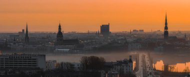 Riga 'nın eski kasabasını aydınlatan sıcak bir gündoğumu, St. Peter Kilisesi, Riga Katedrali, Daugava Nehri üzerinde bir köprü ve aydınlık sabah sisi yer alıyor..