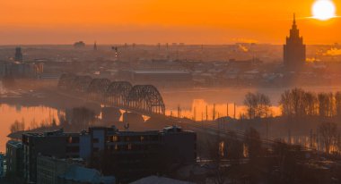 Ilık bir gündoğumu Letonya 'nın Riga kentini aydınlatıyor, arkada Daugava Nehri üzerindeki kemerli demiryolu köprüsü ve Letonya Bilimler Akademisi yer alıyor..