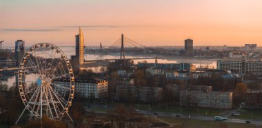 Riga, Letonya 'nın sakin gündoğumu manzarası, Vansu Köprüsü, Z Kuleleri, büyük bir dönme dolap ve sıcak ışığı yansıtan Daugava Nehri..