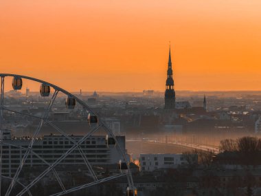 Riga, Letonya üzerinde sakin bir gün doğumu, Aziz Peter Kilisesi kulesi, ön planda bir dönme dolap, ve yumuşak sabah ışığında sıcak turuncu bir gökyüzü..