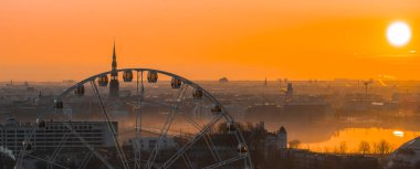 Sıcak bir gündoğumu Riga, Letonya 'yı aydınlatır. Önünde dönme dolap, Aziz Peter Kilisesi kulesi, tarihi binalar ve Daugava Nehri bulunur..