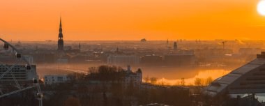 Sıcak bir gündoğumu Riga 'nın eski kasabasını aydınlatıyor. St. Peter Kilisesi kulesi, tarihi çatılar, Daugava Nehri ve Letonya Ulusal Kütüphanesi..