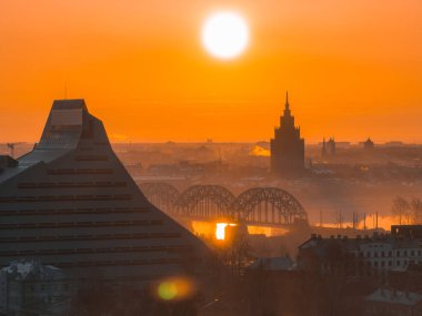 Görüntü Riga, Letonya 'da, Ulusal Kütüphane, Letonya Bilim Akademisi, Daugava Nehri ve bir demiryolu köprüsünün yer aldığı bir güneşin doğuşunu gösteriyor..