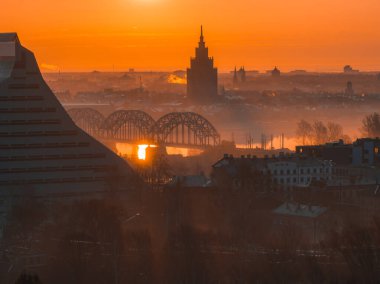 Letonya 'nın başkenti Riga' da sıcak bir gündoğumu, Daugava Nehri, Letonya Bilim Akademisi ve Ulusal Kütüphane üzerindeki kemerli demiryolu köprüsünü aydınlatıyor..