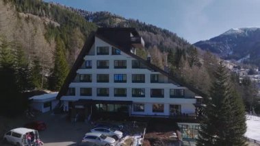 Dolomitler 'de, ormanlar ve kar tozlu yamaçlarla çevrili lüks bir Alp tarzı otelin havadan görünüşü. Kamera düzgünce hareket eder, ayarları gösterir.