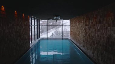 Kapalı havuz alanı, tavandan tavana pencereler, karlı alp manzarası, kayak yamacı, hareket halindeki kar küreme, ahşap paneller ve modern aydınlatma tesisatları..