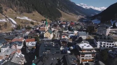 Ischgl, Avusturya 'nın havadan görünüşü, renkli çatıları olan lüks bir otel, yeşil kulesi olan bir kilise, Alp tarzı binalar ve kar kaplı dağları gösteriyor..