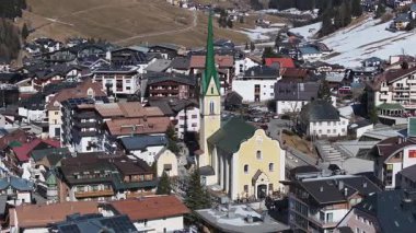 Yeşil kulesi, lüks otelleri, dağlık binaları, ormanlı tepeleri ve kar yamaçları olan sarı bir kilisenin havadan görüntüsü..