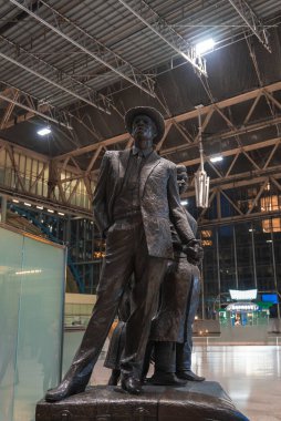 Takım elbiseli ve şapkalı bir adamın bronz heykeli Waterloo tren istasyonunda çelik çerçeveli tavanı, cam duvarları ve görünür tabelaları ile güvenle duruyor..