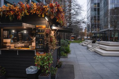 Sonbahar çiçekli çatısı, saksı bitkileri ve menü panosu olan samimi yiyecek tezgahı. Etrafı cam binalar, park bankları ve bulutlu Londra gökyüzü ile çevrili..