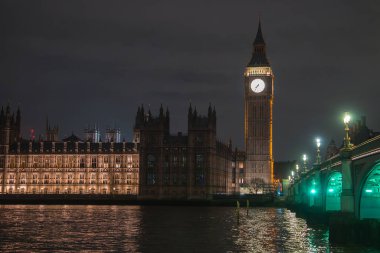 Büyük Ben ve Westminster Sarayı gece gökyüzünde parlarken Westminster Köprüsü 'nün yeşil ışıkları Londra' daki Thames Nehri 'ne yansıyor..