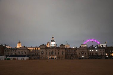 At Muhafızları Yürüyüşü, saat kulesi ile gökyüzüne karşı aydınlık, Londra Gözü sağda pembe parlıyor, ve boş kumlu geçit alanı.