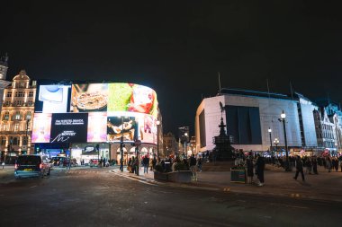 Londra 'daki Piccadilly Circus' un gece manzarasında parlak dijital reklam panoları, Shaftesbury Memorial Fountain ve canlı şehir ortamındaki insanlar.