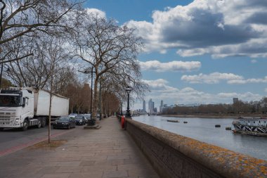 Londra nehir kenarında çıplak ağaçlar, klasik sokak lambaları ve Thames Nehri 'nin olduğu bir yol. Gökdelenler ve demirli bir tekne bulutlu bir gökyüzünün altında görülebilir..