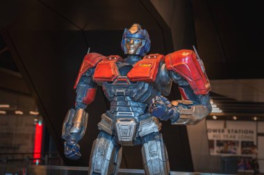 Optimus Prime 'ın kırmızı ve metalik mavi zırhlı detaylı bir heykeli, arka planda modern mimari ve sergi panelleri ile kapalı alanda sergilenmektedir..