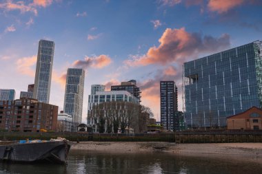 Modern Londra şehrinde cam gökdelenler, geleneksel tuğla binalar ve sıcak turuncu ve pembe bir gökyüzünün altında nehir kıyısında bir mavna bulunur..