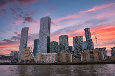 Günbatımında sakin nehir sularına yansıyan cam gökdelenler, konut binaları ve inşaat vinci ile Londra 'nın ufuk çizgisinin görüntüsü.