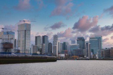 Londra 'daki Canary Wharf' ın modern gökdelenleri Thames Nehri ön planda, inşaat vinçleri ve parçalı bulutlu bir akşam üstü gökyüzü..