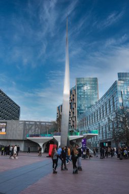 Uzun metalik bir kule, cam binalar, renkli aksanlı yükseltilmiş bir platform ve açık alandan hoşlanan insanlar içeren hareketli bir Londra meydanı..