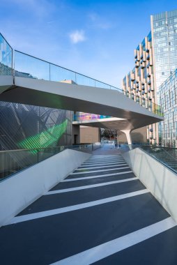 Pürüzsüz, cam parmaklıklı yaya geçidi, Londra 'da açık mavi bir gökyüzünün altında geometrik tasarımları ve yansıtıcı yüzeyleri olan yüksek binalara çıkıyor..