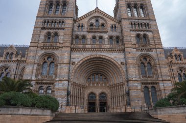 Londra 'daki Doğa Tarihi Müzesi' nin büyük cephesinde Romanca mimari, kemerli pencereler, yüksek kuleler ve ön planda yeşillik yer alıyor..