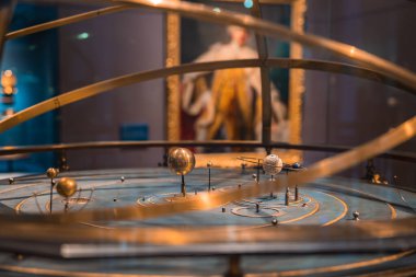 Londra müzesinde altın yüzükler ve mavi tabanlı ayrıntılı bir orrery. Altın çerçevede bulanık bir kraliyet portresi arka planda görünür..
