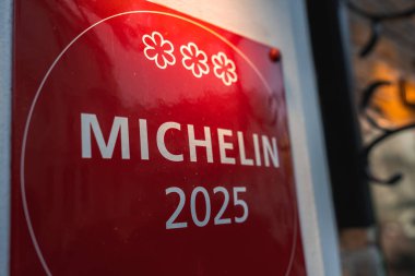 Üç yıldızlı kırmızı Michelin 2025 plaketi açık renkli bir duvara monte edilmiştir. Bir dekoratif işlenmiş demir özelliği kısmen sağda görülebilir..
