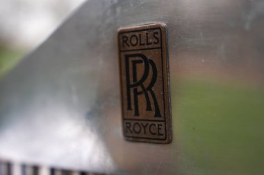 Rolls Royce ambleminin RR logosu ve oymalı plaka ile detaylı bir görüntüsü. Bulanık arkaplan açık hava ortamında yeşillik belirtileri gösteriyor.