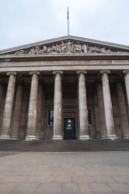 British Museum 'un Londra' daki neoklasik cephesinde uzun flütlü sütunlar, heykelleri olan üçgen bir soy ağacı ve bayrak direğinde bir İngiliz bayrağı var..