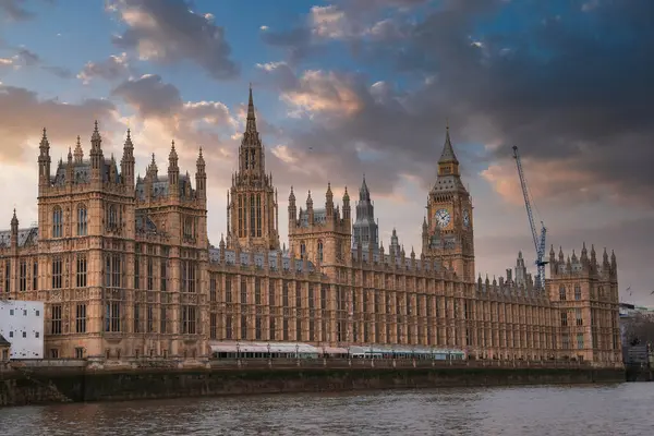 İngiltere, Londra 'daki Westminster Sarayı ve Big Ben Sarayı' nda Gotik Diriliş mimarisi, dramatik bir gökyüzü ve Thames Nehri ön planda..