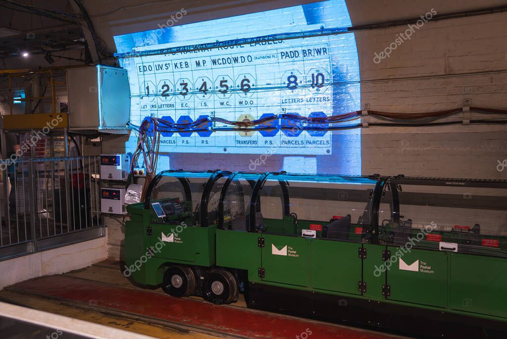 A green train with transparent covers on the Mail Rail tracks in London. A projection shows a vintage postal sorting board in an industrial setting.