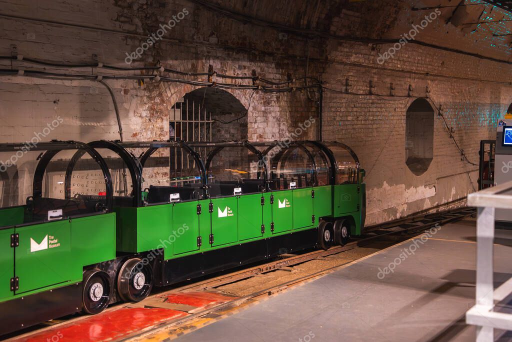 A green miniature train with transparent domed roofs sits on tracks in a brick lined underground tunnel, part of the historic Mail Rail in London.