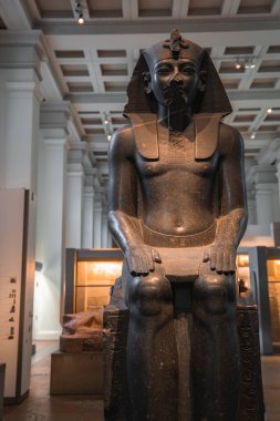 Tahtta oturan Mısır firavununun koyu renkli taş heykeli Londra 'daki bir müzede sergileniyor. Arka planda yüksek tavanlar, sütunlar ve vitrinler var..