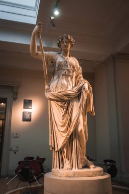 Londra müzesinde yüksek tavanlı ve tabelalı yumuşak ışıklandırmanın altında bir asayı tutan kadın figürünün mermer heykeli..