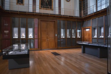 Zarif bir müze galerisi. Parlatılmış ahşap zeminler, objeli cam vitrinler, merkezi ahşap bir kapı ve ikinci bir balkon..