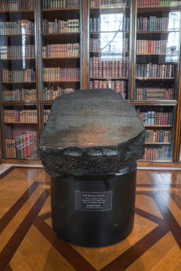Rosetta Taşı, kitaplıklar ve ahşap zeminlerle çevrili, muhtemelen İngiliz Müzesi 'nde bulunan bilgilendirici bir plaketle siyah bir kaideye dayanır..
