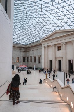 Londra 'daki British Museum' un Büyük Mahkemesi 'nde cam ve çelik bir çatı, neoklasik bir yapı ve açık alanda yürüyen ve toplanan ziyaretçiler yer alıyor..