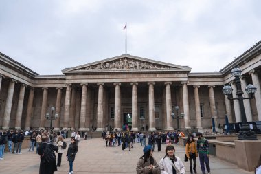 Londra 'daki British Museum' da uzun iyonik sütunları, üçgen pedikürleri ve Union Jack bayrağı bulunan neoklasik mimari sergilenmektedir. Ziyaretçiler avluyu dolduruyor.